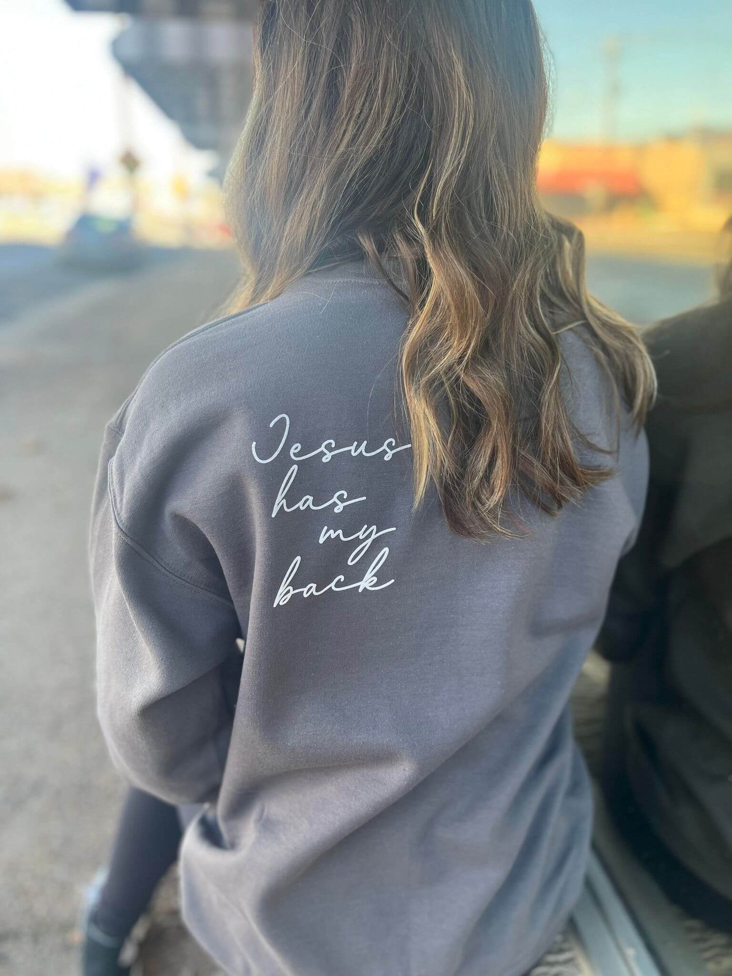 Woman wearing a charcoal sweatshirt with "Jesus has my back" printed on the back, showcasing comfort and style.
