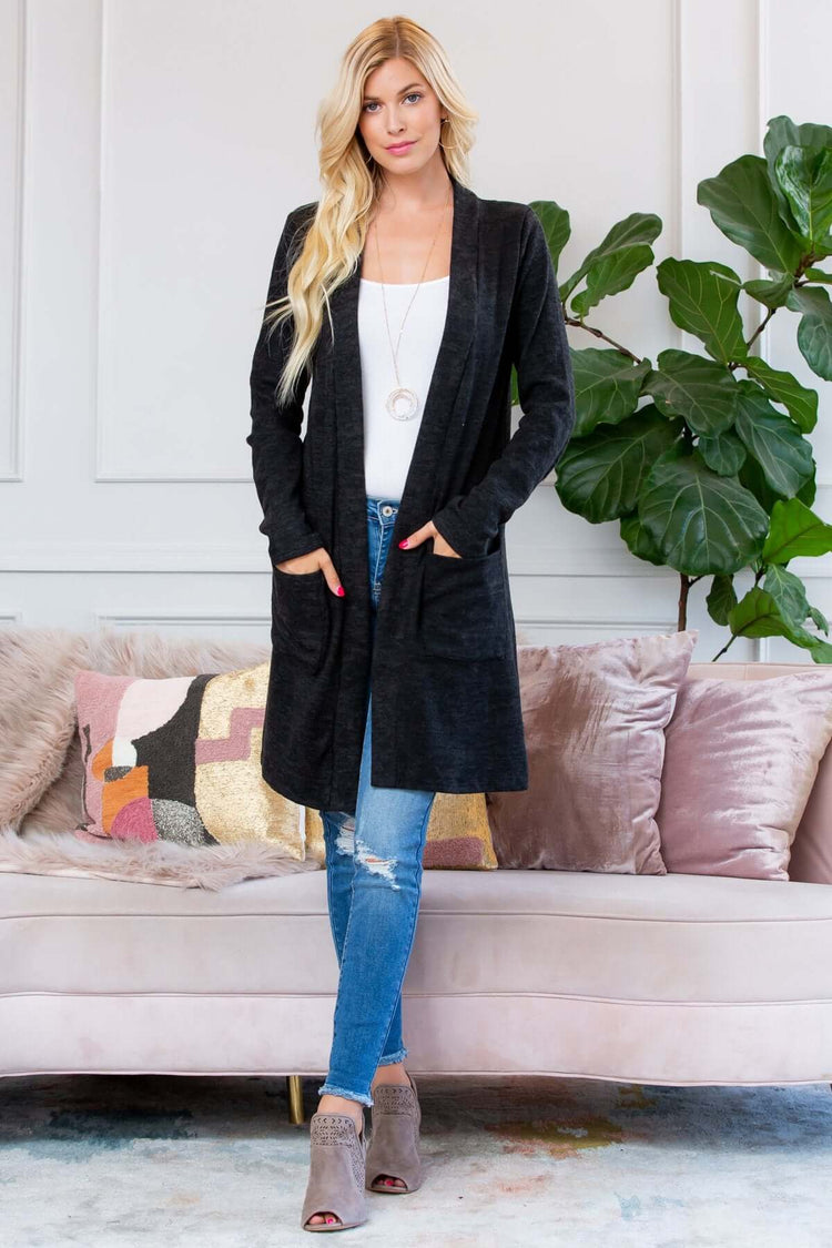 Model wearing a black brushed sweater pocket cardigan over a white top and blue jeans, standing in a cozy living room.