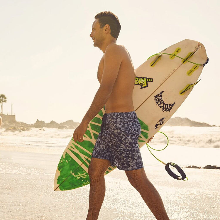 Man walking on the beach in Sustainable Surf Geometric Gray boardshorts, holding a surfboard.