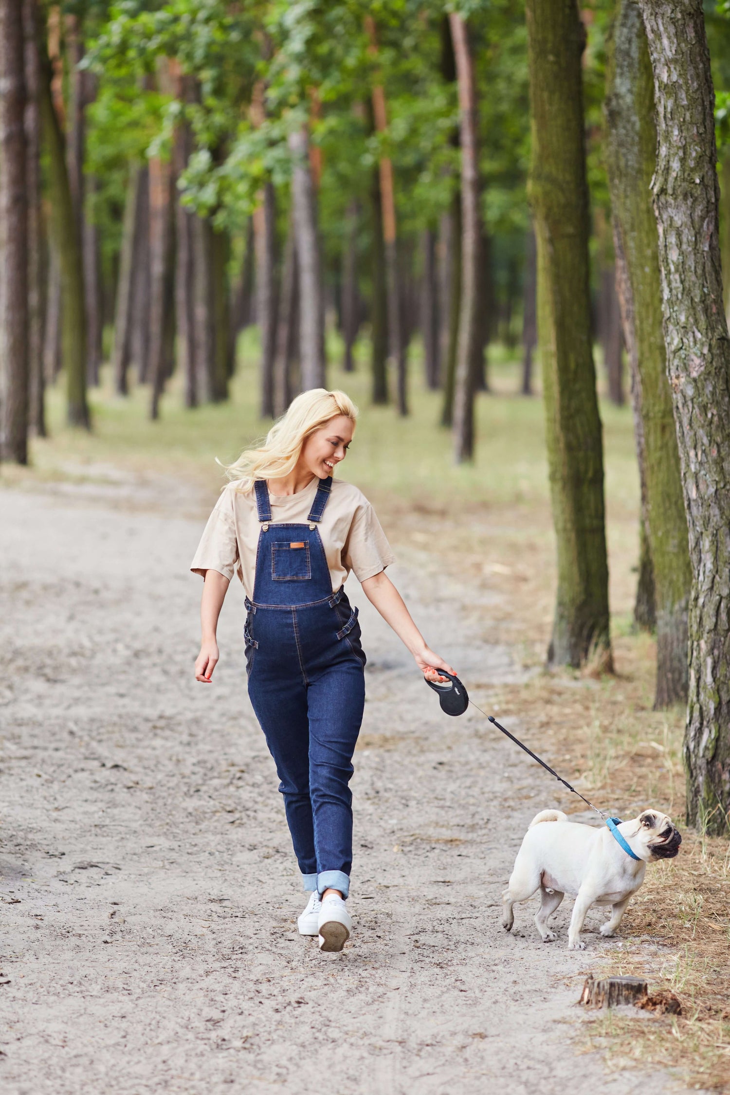 Woman in denim overalls walking a dog on a forest path, enjoying a casual outdoor day.