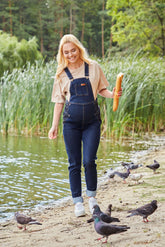 Happy woman wearing belly support maternity overalls by the water, enjoying a casual day while feeding birds.