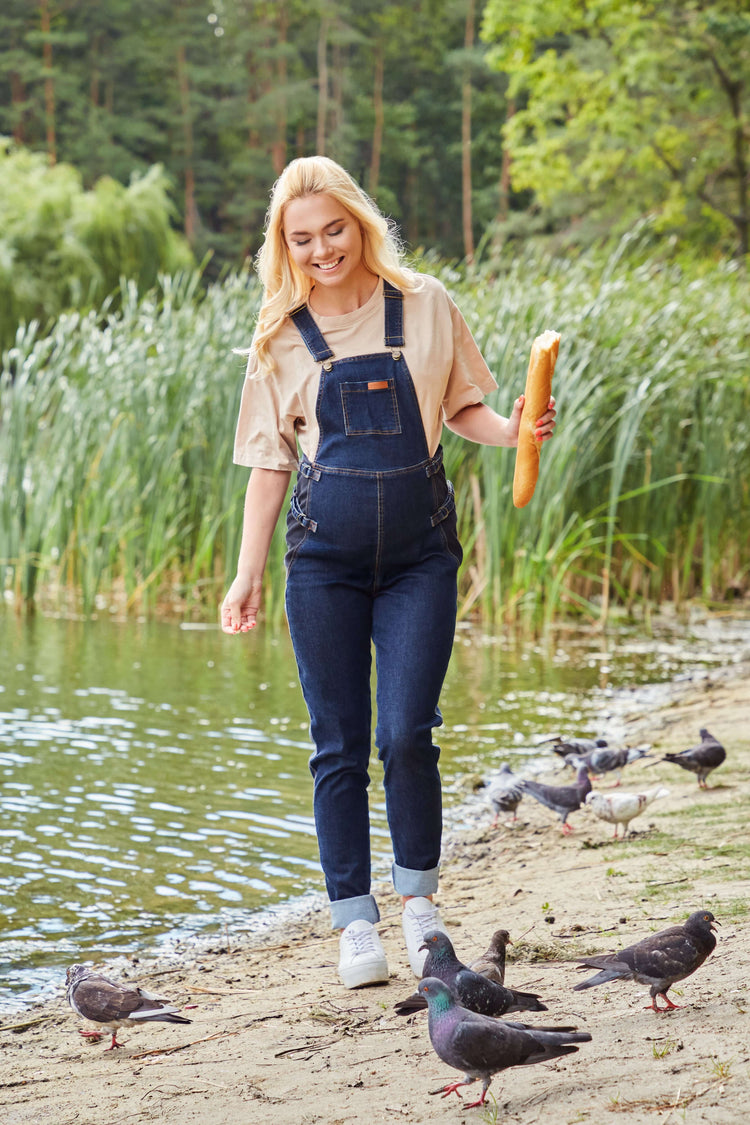 Happy woman wearing belly support maternity overalls by the water, enjoying a casual day while feeding birds.