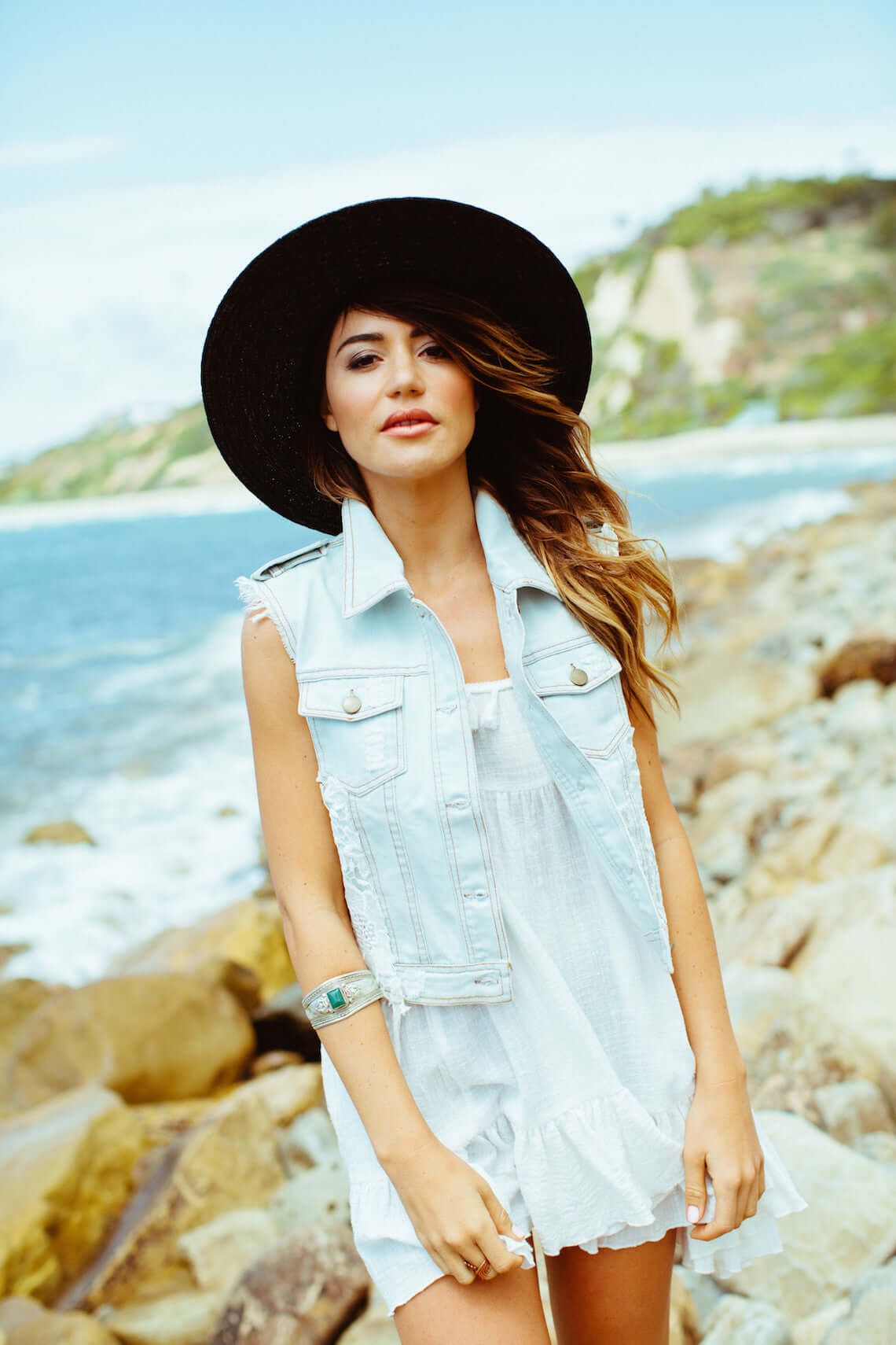 Model wearing Margot Vest, a denim vest with lace back, by the beach, showcasing a stylish summer look.