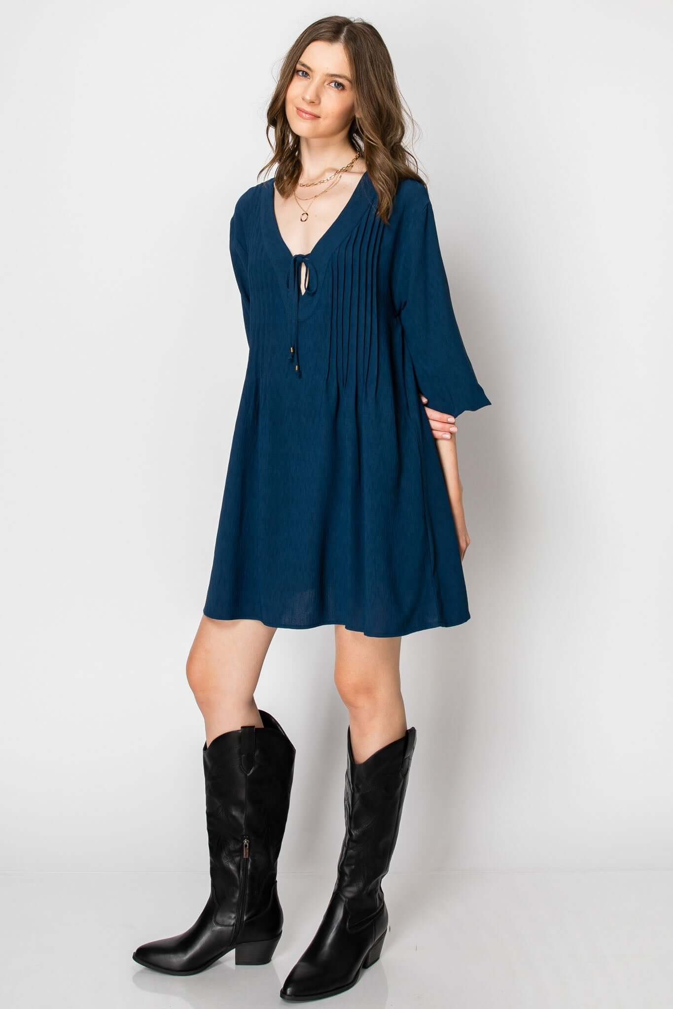 Long sleeve flowy A-line dress in teal with pintuck details, front tie, and pockets, styled with black western boots.