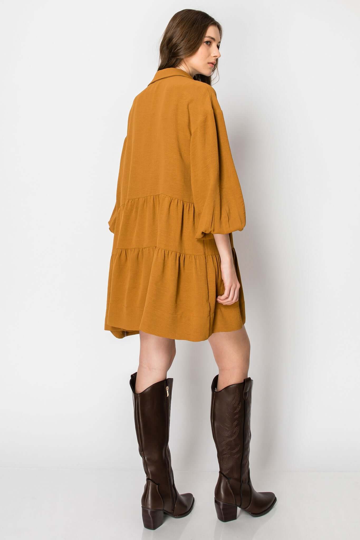 Model wearing a long sleeve collared tiered ruffle dress in mustard, styled with tall brown boots for a casual fall look.