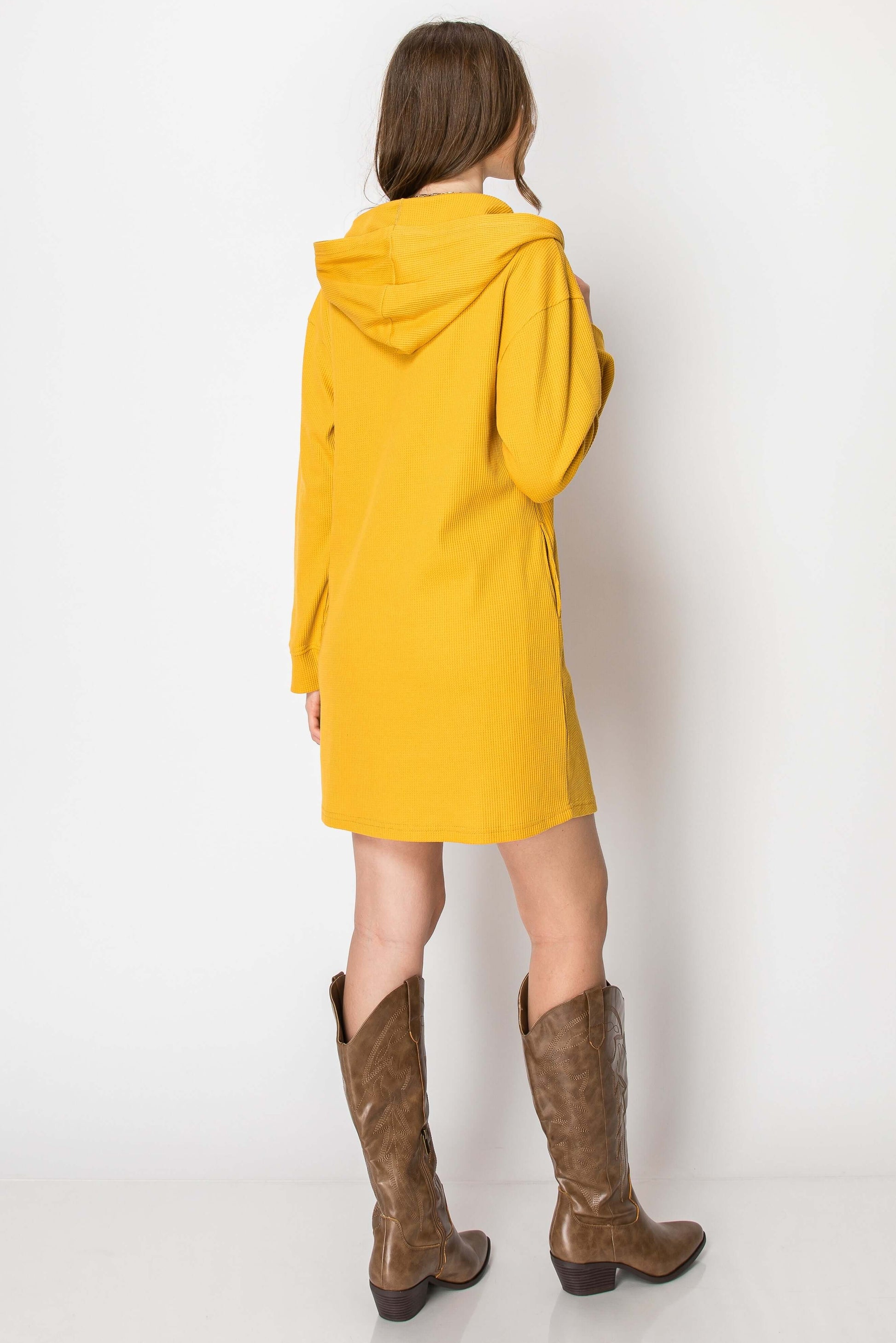 Back view of a yellow waffle knit long sleeve hoodie dress with pockets, styled with brown knee-high boots.