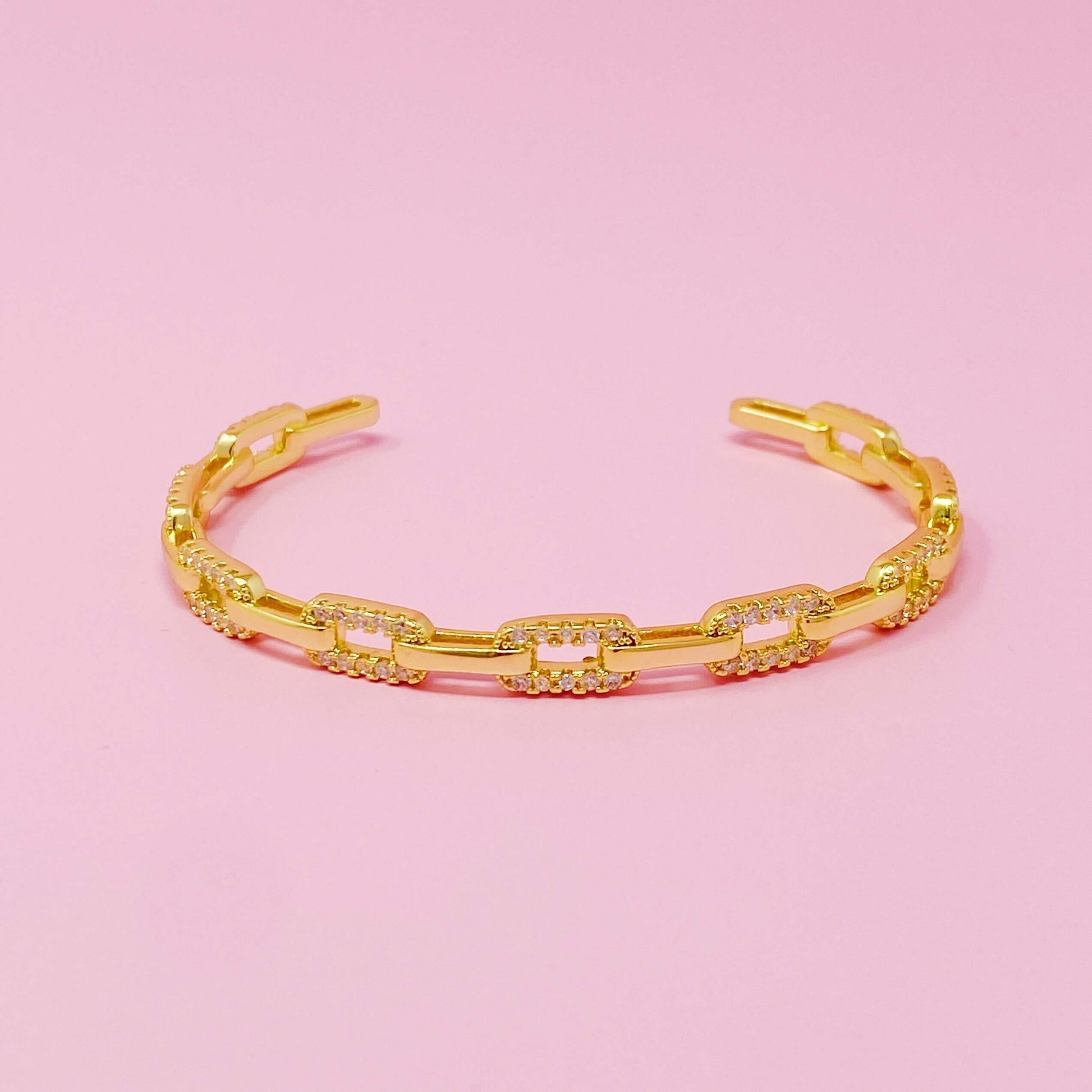 Stylish alternatively linked open bangle bracelet in 18k gold plated brass with cubic zirconia on pink background.