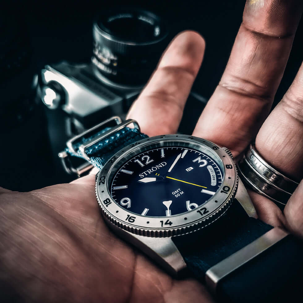 Stylish 24h GMT watch in hand with blue dial, stainless steel case, and NATO strap, capturing a classic look.