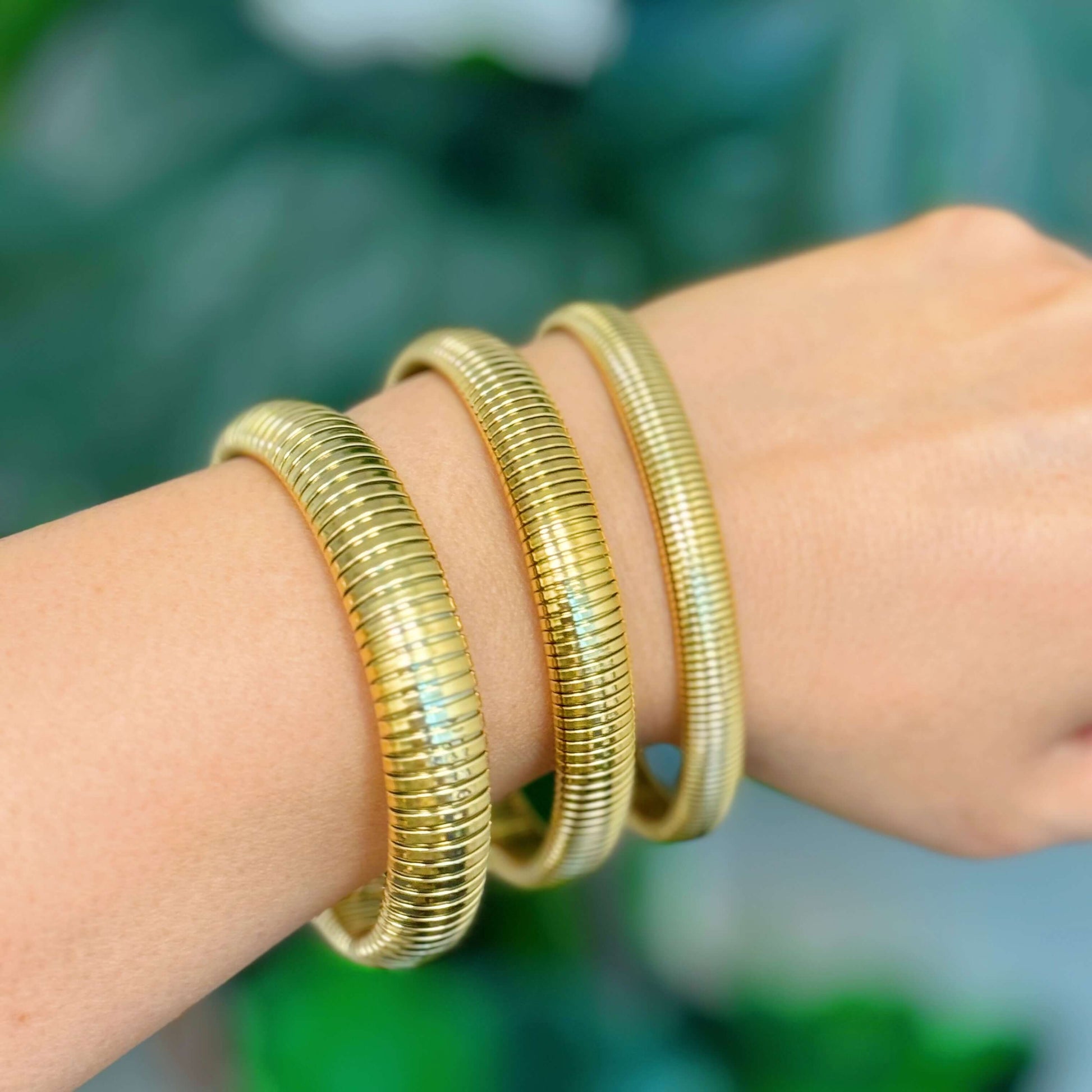 Close-up of three golden spring soft bangle bracelets worn on a wrist against a blurred green background.