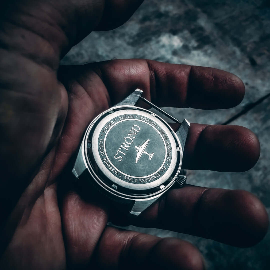 Hand holding a stainless steel watch with engraved back showcasing the brand STROND.