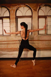 Dancer wearing a black jumpsuit, posing gracefully in an empty studio with large windows.