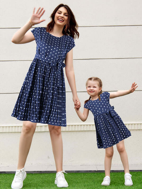 Mother and daughter wearing matching woven polka dot pleated dresses, smiling and holding hands outdoors.