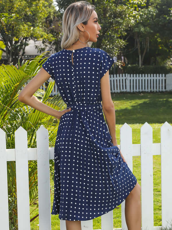 Rear view of a woman in a navy polka dot pleated midi dress, showcasing its high waist and half sleeves.