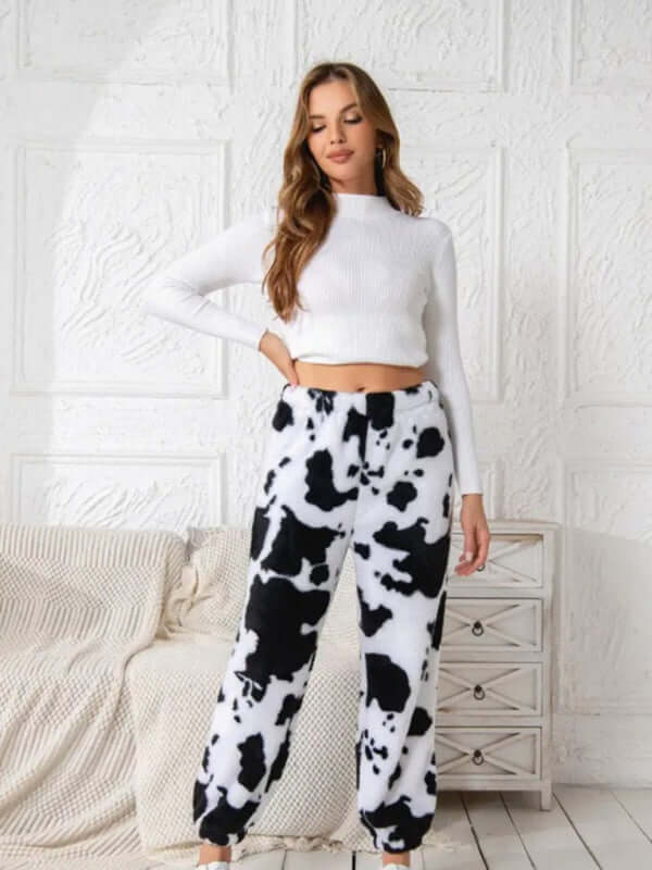 Model wearing cow print fleece pants and a white crop top in a cozy indoor setting.